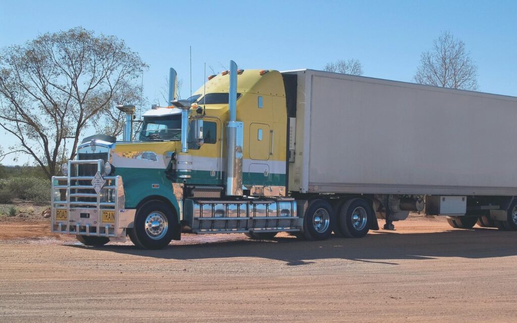Blue and yellow semi-truck with a white box trailer parked on a dirt clearing.