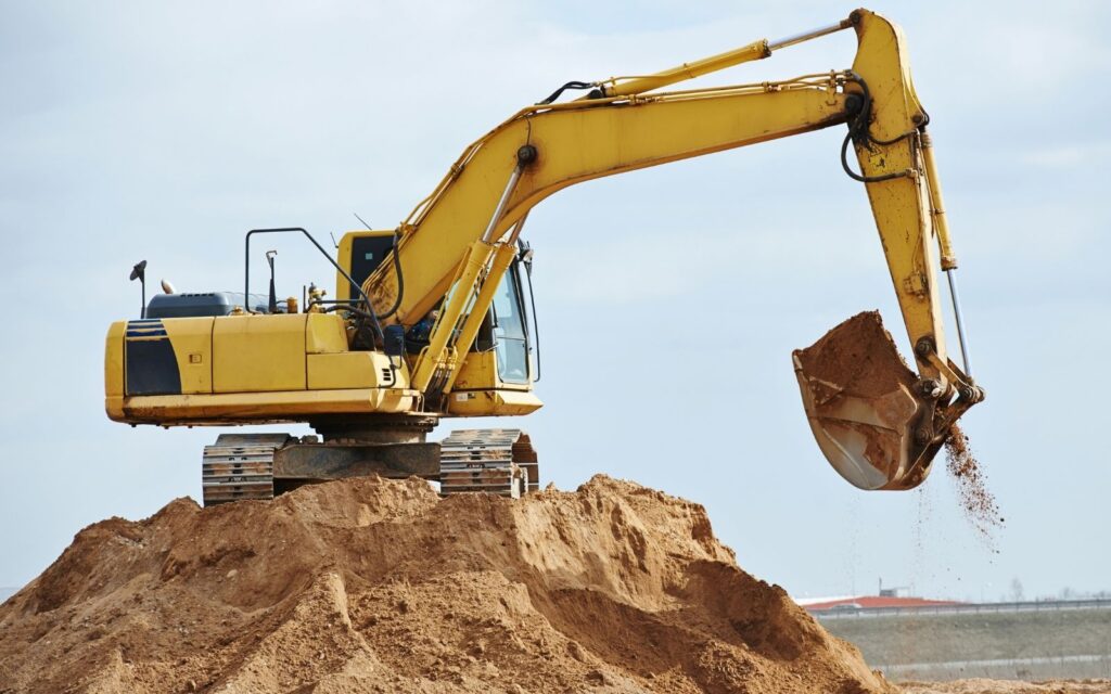 Yellow tracked excavator digging from the top of a soil mound.